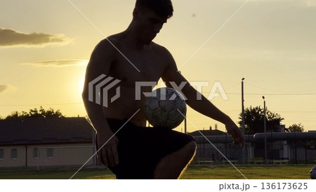 Young man juggling soccer ball on stadium at sunset. Professional footballer kicking ball at green field. Sportsman practicing tricks at meadow with sunlight at background. Freestyle football 136173625