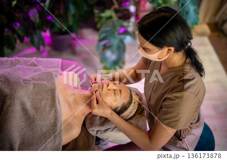 Tranquil woman lying on spa bed getting anti aging lifting special facial massage from cosmetologist 136173878
