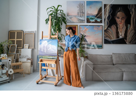 Pleased happy woman artist posing with finished artwork on easel in modern loft apartment workshop 136173880