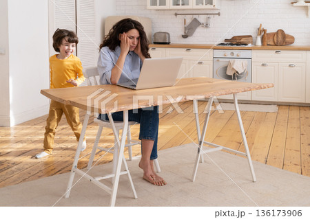 Irritated mother remote work from home in covid-19 pandemic with naughty son running in kitchen Irritated mother remote work from home in covid-19 pandemic with naughty son running in kitchen 136173906