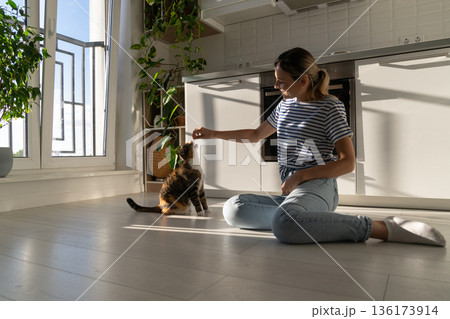 Good-natured Caucasian woman plays with kitten and has good time sits on kitchen floor near stove 136173914