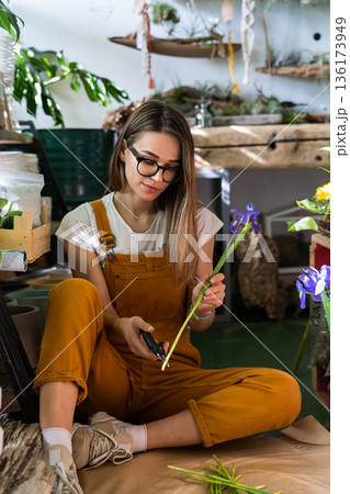 Florist woman cutting stem of irises flowers using secateurs, sitting on the floor in flower store. Florist woman cutting stem of irises flowers using secateurs, sitting on the floor in flower store. 136173949