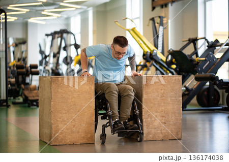 Confident man athlete in wheelchair doing push-ups strengthening exercise on wooden cubes in gym. 136174038