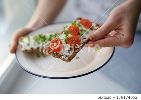 Hands holding plate with rye crisp bread with creamy cheese tofu, cherry tomato and micro greens 136174052