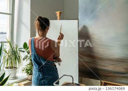 Focused female artist painting picture at home studio, holding paintbrush and looking at canvas. Focused female artist painting picture at home studio, holding paintbrush and looking at canvas. 136174069