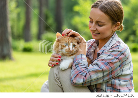 Smiling woman in checked shirt hugging and embracing with tenderness and love domestic ginger cat, stroking on the head, outdoors in sunny day. Love to the animals Smiling woman in checked shirt hugging and embracing with tenderness and love domestic ginger cat, stroking on the head, outdoors in sunny day. Love to the animals 136174110