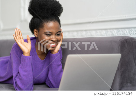 Hello nice meet you. Happy Afro-American woman with afro hairstyle lying on couch, waving hi greeting gesture, looking at camera webcam, say hello, talking on video chat with friends on laptop. Hello nice meet you. Happy Afro-American woman with afro hairstyle lying on couch, waving hi greeting gesture, looking at camera webcam, say hello, talking on video chat with friends on laptop. 136174154