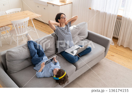 Woman resting after work from home office on laptop sit on couch child playing on tablet. Lockdown. 136174155