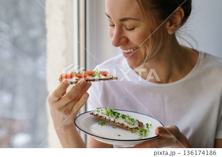 Woman eating rye crisp bread with creamy vegetarian cheese tofu, tomato, micro greens. Healthy food 136174186