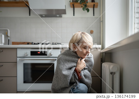 Woman wrapped in knitted plaid sitting near central heating radiator temperature, cold at home 136174199