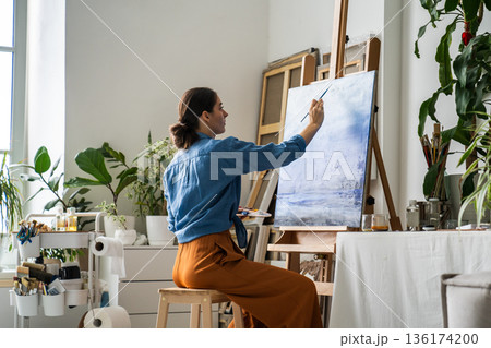 Relaxed woman artist painting picture on easel at home, sitting on chair in cozy creative atmosphere 136174200