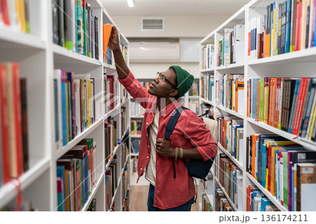 Nerd student african american man choosing book in university library taking it from shelf. Nerd student african american man choosing book in university library taking it from shelf. 136174211