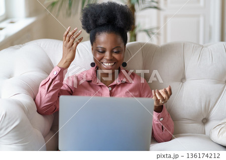 African young woman sitting on couch talking on video chat online or webinar with friends at home. 136174212