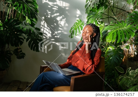 Dreamy African American woman with closed eyes relaxing in home garden filled with tropical plants. Dreamy African American woman with closed eyes relaxing in home garden filled with tropical plants. 136174257