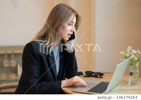 Smiling businesswoman working on laptop and talking on smartphone during lunch break in cafe. 136174263