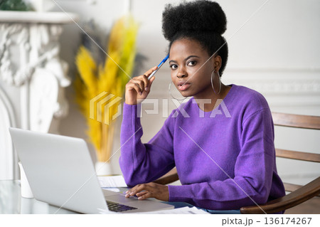 Afro-American millennial student woman with afro hairstyle looking at camera while making researches browsing information on laptop, preparing for exam online, holding a pen. 136174267