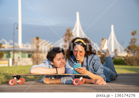Trendy couple browsing web on smartphone outdoors using 4g internet connection lying on longboard 136174276