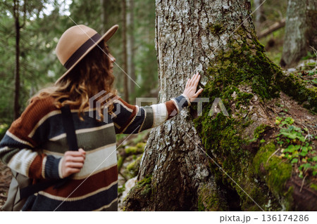 Female tourist with backpack and travel equipment touching tree while standing in the forest. 136174286
