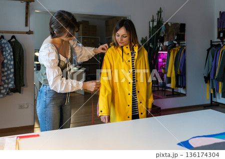 Fashion designer workshop with female tailor measuring and fitting bespoke yellow rain jacket. 136174304