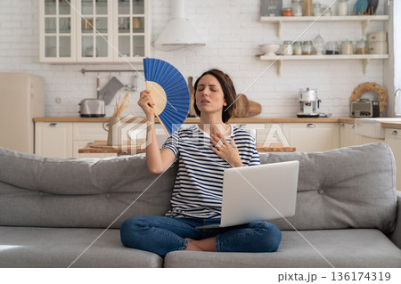 Young woman suffers from heatstroke flat without air-conditioner waving fan sitting on couch at home 136174319