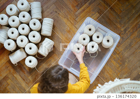 Top view of woman freelancer holding white macrame cotton ropes in skeins, puts working material in a plastic box. Crochet, weaving crafts Top view of woman freelancer holding white macrame cotton ropes in skeins, puts working material in a plastic box. Crochet, weaving crafts 136174320
