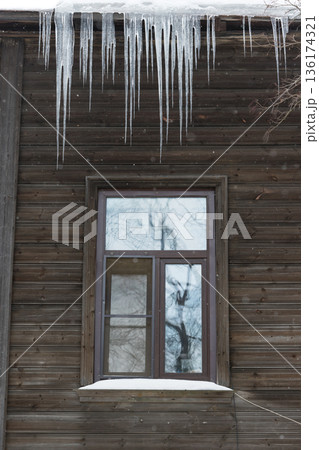 Ice stalactite hanging from the roof with wooden wall. Poor thermal insulation of the roof. Winter season 136174321