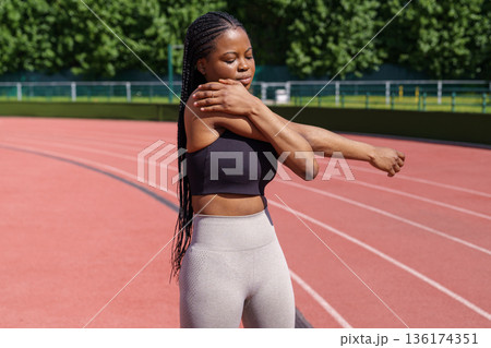 Black young woman with long braids stretches hands and shoulders standing on red track on stadium Black young woman with long braids stretches hands and shoulders standing on red track on stadium 136174351