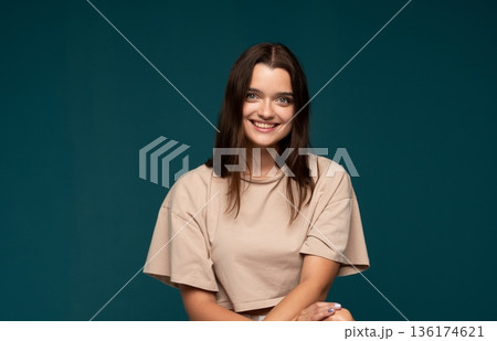 Smiling young woman with natural makeup and brown hair posing against dark green studio background with relaxed and friendly expression Smiling young woman with natural makeup and brown hair posing against dark green studio background with relaxed and friendly expression 136174621