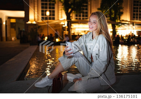 Young woman smiling using smartphone sitting by Vienna fountain 136175101
