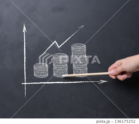 A person's hand with a pencil points to a chalked growth chart with coin-shaped bars on a black chalkboard 136175252