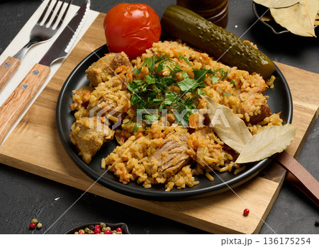 Pilaf with pieces of meat and herbs served on a black plate with pickled cucumber, marinated tomato on a black background 136175254