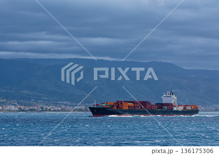 Large cargo vessel passing close to mountainous coastline creating commercial maritime transport scene. 136175306