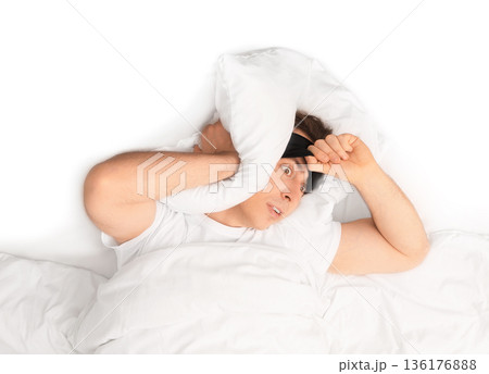 Top view of sleepless man covering ear with pillow. Annoyed guy suffering from insomnia or loud noise lying in white bed with sleep mask. High quality photo 136176888