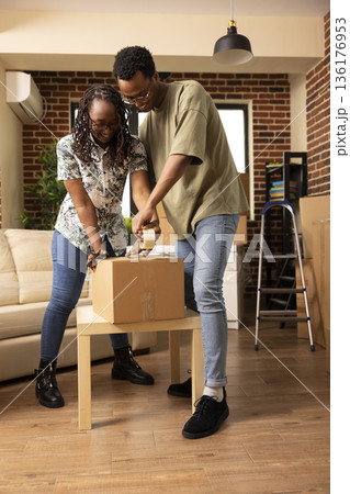 Young married couple packing parcels together in cozy living room. African american husband and wife organizing belongings in brick wall apartment, taping cartons on table with teamwork. Young married couple packing parcels together in cozy living room. African american husband and wife organizing belongings in brick wall apartment, taping cartons on table with teamwork. 136176953
