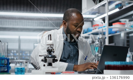 Molecular biologist in lab analyzing batch of blood samples for red blood cell abnormalities. African american man doing research on sanguine fluid vials to diagnose anemia types, camera B 136176990