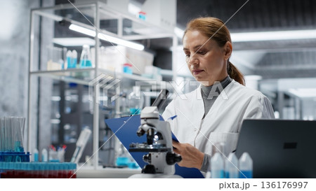 Hematology specialist in lab conducting research on blood sample, taking notes. Epidemiologist in laboratory analyzing sanguine fluid specimen in vial, writing down findings, camera B 136176997