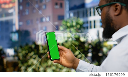 Vertical green screen phone held by black man on a busy city center street, walking around the metropolitan downtown area with skyscrapers and dynamic scenery. Vibrant avenue. Camera B. 136176999