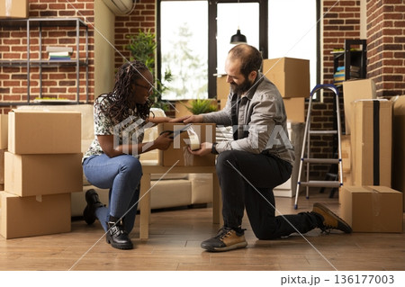 African american girlfriend and caucasian boyfriend kneeling on floor, taping cartons. Happy diverse couple preparing to move out, carefully sealing packages and parcels in bright brick wall room. African american girlfriend and caucasian boyfriend kneeling on floor, taping cartons. Happy diverse couple preparing to move out, carefully sealing packages and parcels in bright brick wall room. 136177003