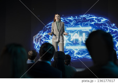 Female business speaker leads futuristic AI-driven presentation with AI graphics, neural networks and deep learning concepts. Business discussion with audience in a high tech auditorium. Female business speaker leads futuristic AI-driven presentation with AI graphics, neural networks and deep learning concepts. Business discussion with audience in a high tech auditorium. 136177017