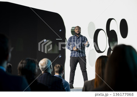 Black chief product officer introducing new smartphone with modern interface and design for marketing announcement. Mixed age audience during launch event focused on phone release. Black chief product officer introducing new smartphone with modern interface and design for marketing announcement. Mixed age audience during launch event focused on phone release. 136177068