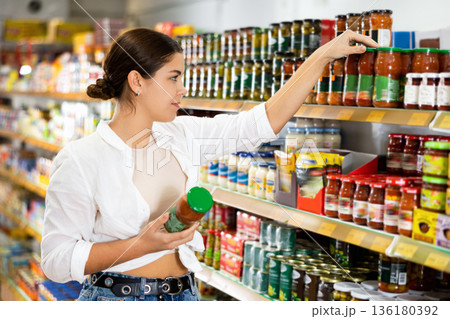 Pensive young girl choosing canned goods in supermarket Pensive young girl choosing canned goods in supermarket 136180392