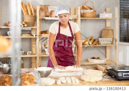 Young woman works as baker, mold baguette from dough, creates long loafs from dough. Young woman works as baker, mold baguette from dough, creates long loafs from dough. 136180413