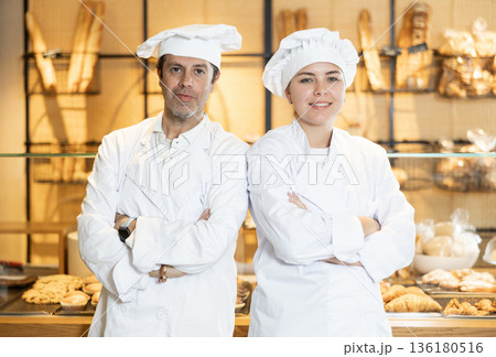 Two positive confident middle-aged male and young female bakery owners in chef jackets standing with crossed hands on chest in bakeshop 136180516