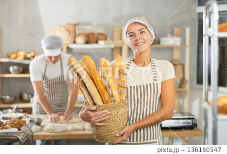 Young female baker holding baguettes in round wicker basket Young female baker holding baguettes in round wicker basket 136180547
