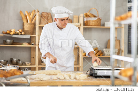 Young male baker cutting and weighing dough on scales in bakehouse 136181118