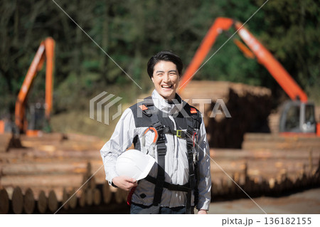 建築現場などでショベルカーを背景にヘルメットをつけた林業など高所作業などのハーネス(安全帯)の男性 136182155