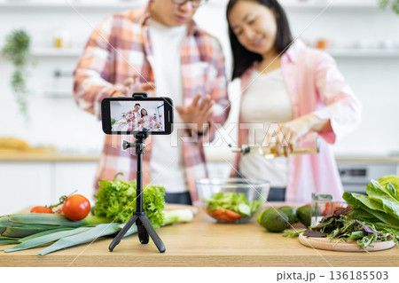 A couple is filming a cooking tutorial in their kitchen, preparing a fresh salad with vegetables and oil A couple is filming a cooking tutorial in their kitchen, preparing a fresh salad with vegetables and oil 136185503