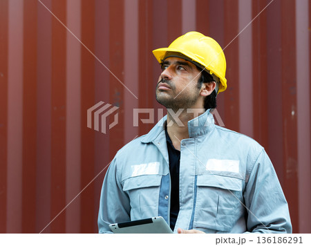 Industrial worker wearing safety helmet and protective jacket holding a tablet, standing in front of cargo container, representing construction management, logistics inspection, safety control, and Industrial worker wearing safety helmet and protective jacket holding a tablet, standing in front of cargo container, representing construction management, logistics inspection, safety control, and 136186291