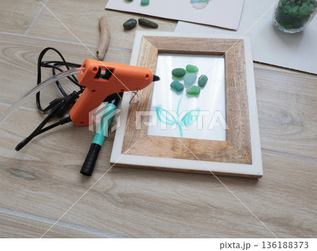 Sea glass flower in frame. Making art from natural materials. Glue gun and driftwood for picture. Hobbies and creativity. Reuse and nature, ecology. Sea glass flower in frame. Making art from natural materials. Glue gun and driftwood for picture. Hobbies and creativity. Reuse and nature, ecology. 136188373