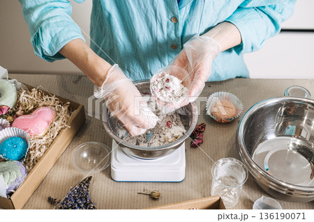 Hands mix white powder and dried botanicals in metal bowl on digital scale surrounded by finished products. Handmade bath bombs process, cosmetic chemistry at home, artisan soap making. 136190071